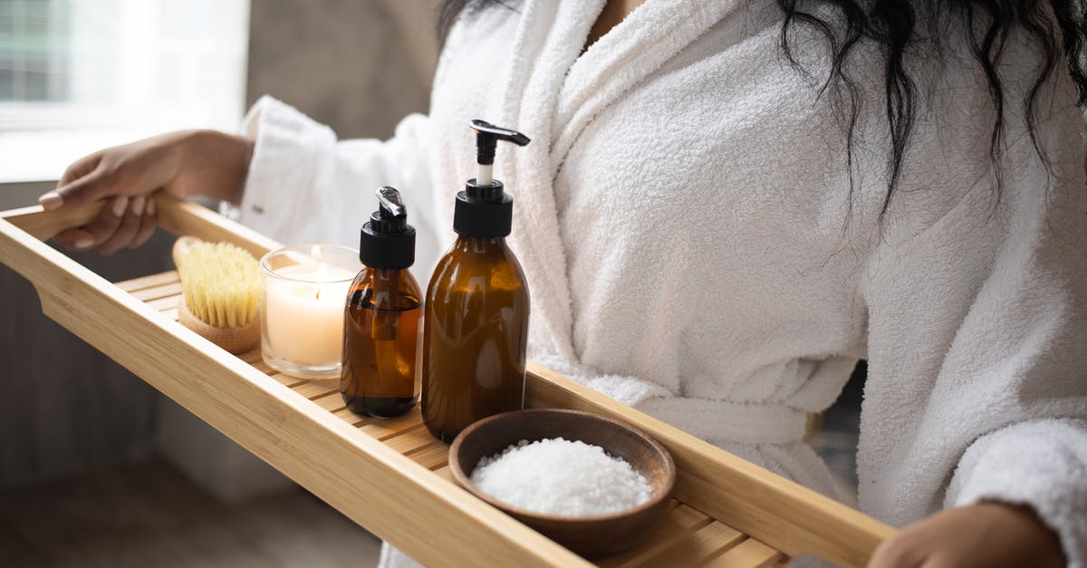 Adding salt to the oil making potato chips - Unrecognizable African American female in white bathrobe with bottles of cosmetic products salt and burning candle on wooden tray in hands