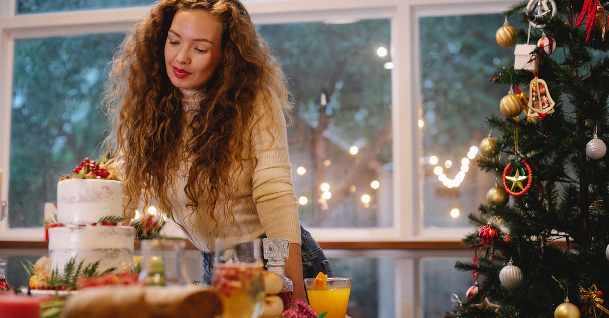 Advice on two-tier cake and height - Low angle of serious female leaning on served festive table with two tier cake near decorated Christmas tree Advice on two-tier cake and height - Low angle of serious female leaning on served festive table with two tier cake near decorated Christmas tree