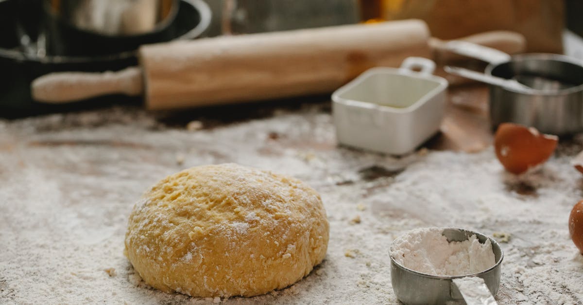 After rolling pizza dough, how do I move it to a pan without ruining it? - Ball of raw dough placed on table sprinkled with flour near rolling pin dishware and measuring cup in kitchen on blurred background