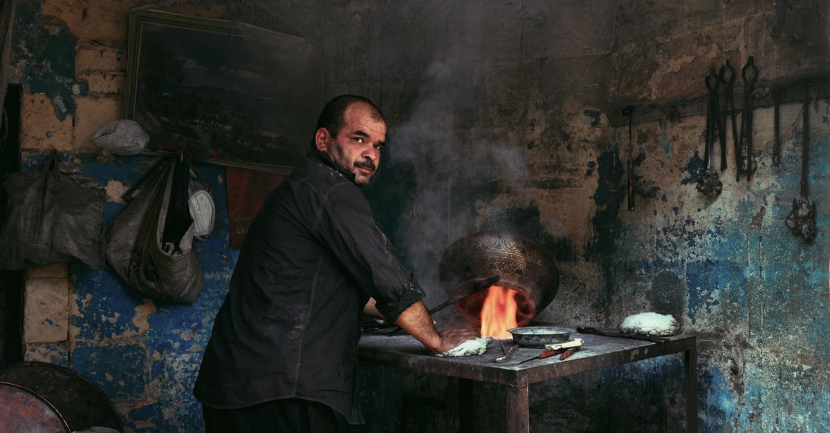 An electronic tool for mixing while the pot is on the fire - Back view of ethnic worker heating pot with burning flame while looking at camera in smithy