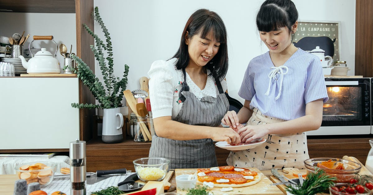Baking pizza without an oven - Happy smiling Asian mother and daughter in aprons putting ingredients on dough while cooking pizza together in kitchen Baking pizza without an oven - Happy smiling Asian mother and daughter in aprons putting ingredients on dough while cooking pizza together in kitchen