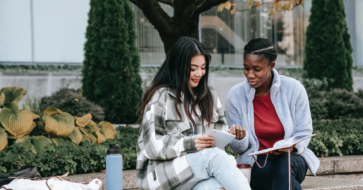 Best practices for using taps while avoiding cross contamination - Smiling multiethnic girlfriends talking and sharing smartphone on street bench Best practices for using taps while avoiding cross contamination - Smiling multiethnic girlfriends talking and sharing smartphone on street bench