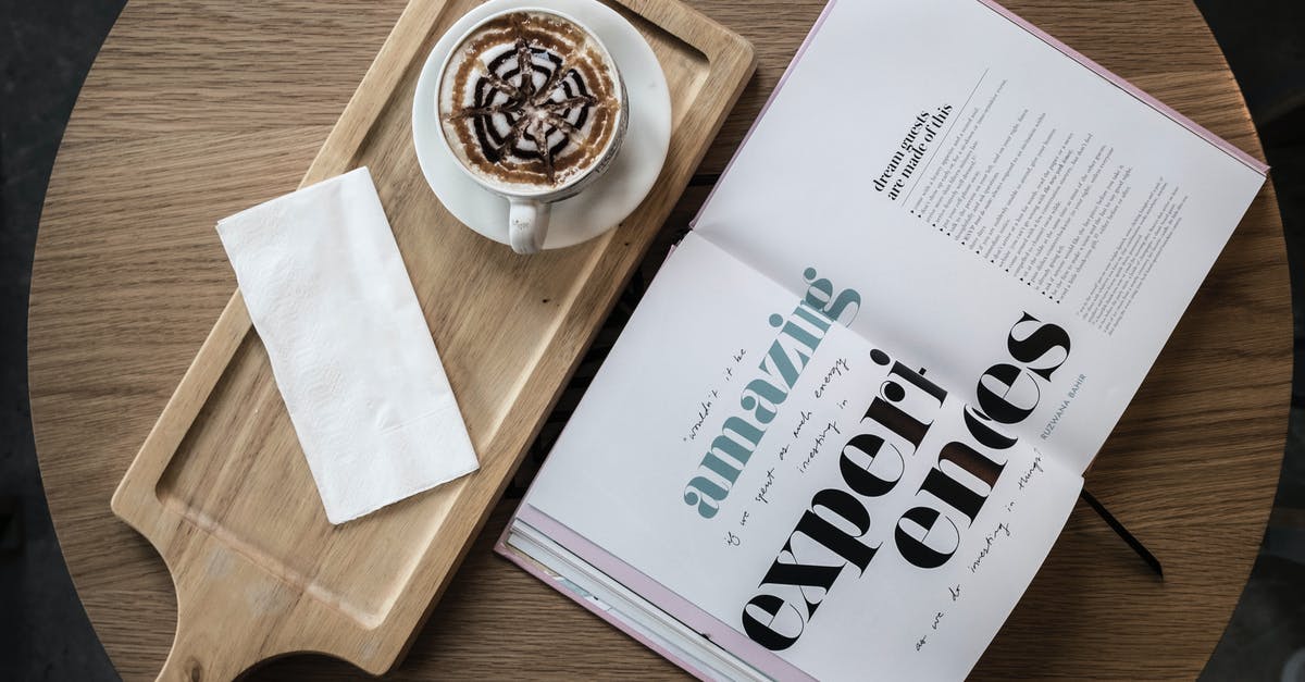 breaking off chocolate from a block of chocolate - Top view of cup of cappuccino garnished with chocolate topping served with saucer and napkin on wooden board placed on cafe round table with open book with amazing experiences title on pages
