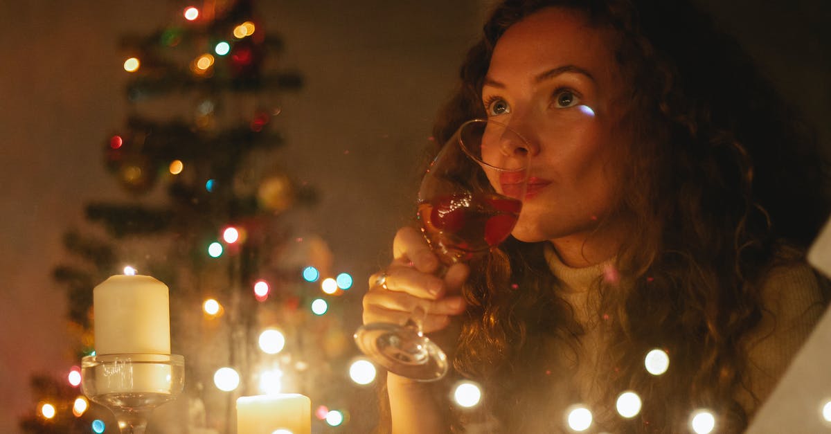 Cooking away alcohol - Through glass of dreamy female with glass of champagne sitting among shiny garlands and burning candles during Christmas celebration Cooking away alcohol - Through glass of dreamy female with glass of champagne sitting among shiny garlands and burning candles during Christmas celebration