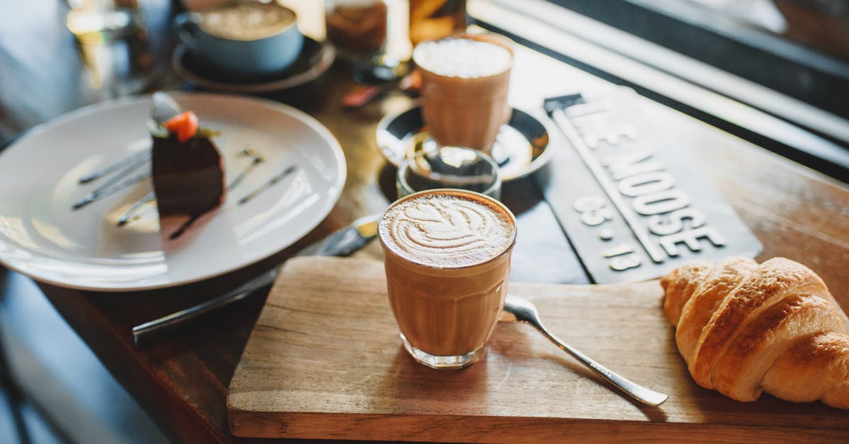 Dalgona / whipped coffee without sugar? - From above of glass of cold coffee drink served with fresh crunchy croissant on wooden board near plate with chocolate cake in cozy cafe