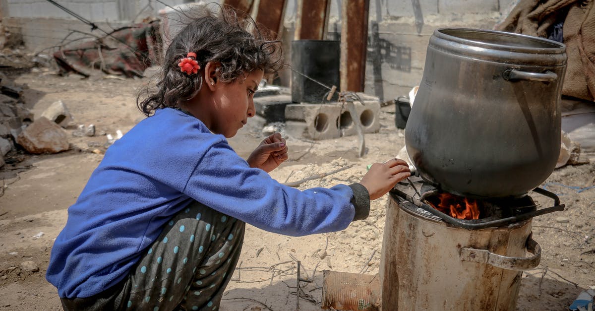 Damage to pan bottom - Side view of ethnic kid sitting near furnace with metal pan placed on fire in daylight in poor destroyed district Damage to pan bottom - Side view of ethnic kid sitting near furnace with metal pan placed on fire in daylight in poor destroyed district