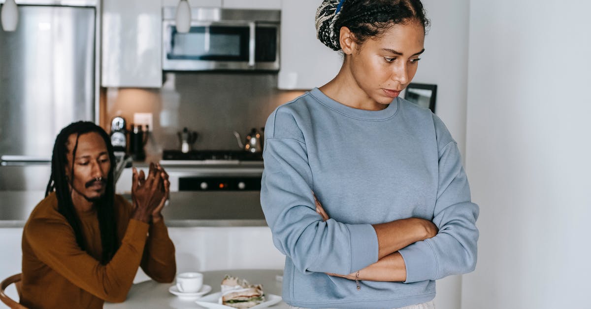Dense and gluey centered cake problem - Young discontented African American female with folded arms against male partner at kitchen table during quarrel Dense and gluey centered cake problem - Young discontented African American female with folded arms against male partner at kitchen table during quarrel
