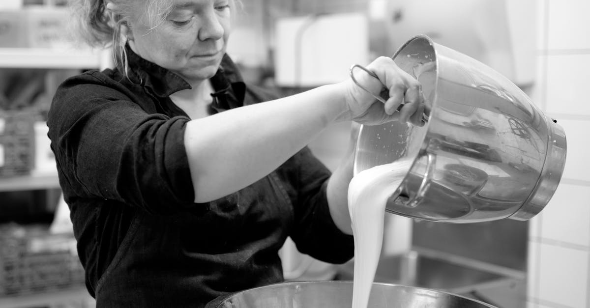 Fatty liquid vs non fatty liquid in dough - Black and white of focused adult female chef in black wear with hair tied up pouring white liquid eatable mass in big glistening metallic dish Fatty liquid vs non fatty liquid in dough - Black and white of focused adult female chef in black wear with hair tied up pouring white liquid eatable mass in big glistening metallic dish