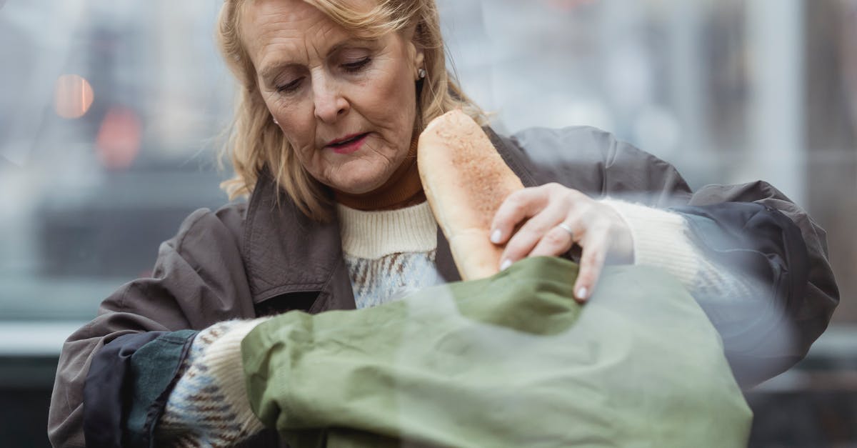 Flies in my bag of bread - bad bread? - Through glass view of senior female with wrinkled skin in warm clothes putting long loaf in textile bag in daytime