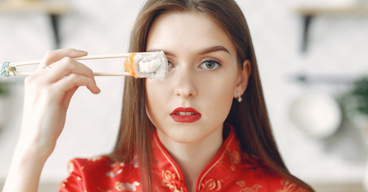 How to dress sushi to better enhance all its flavours? - Young lady with dark hair in stylish wear holding chopsticks with sushi and covering eye while resting in light kitchen at home