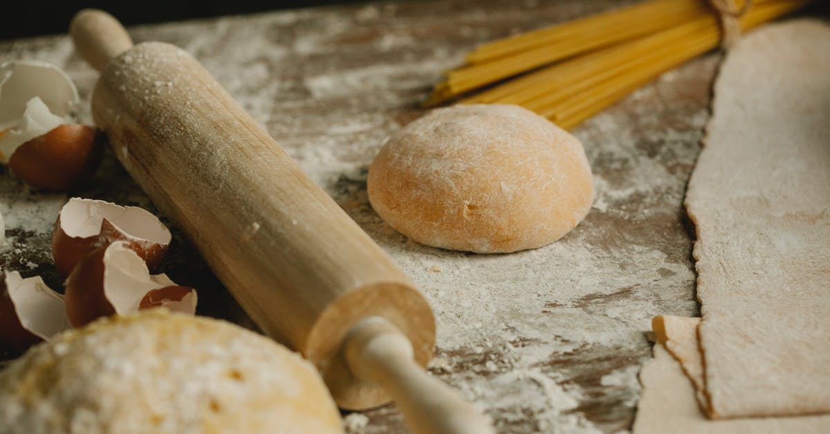 How to layer a Lasagne - Uncooked balls of dough near rolling pin placed on wooden table sprinkled with flour with eggshell and raw spaghetti in kitchen