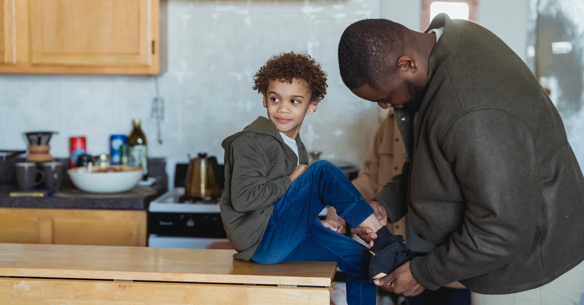 I put my cheddar in my kitchen cabinet - Focused African American father helping cheerful black son to put on footwear while sitting on wooden table in modern kitchen I put my cheddar in my kitchen cabinet - Focused African American father helping cheerful black son to put on footwear while sitting on wooden table in modern kitchen