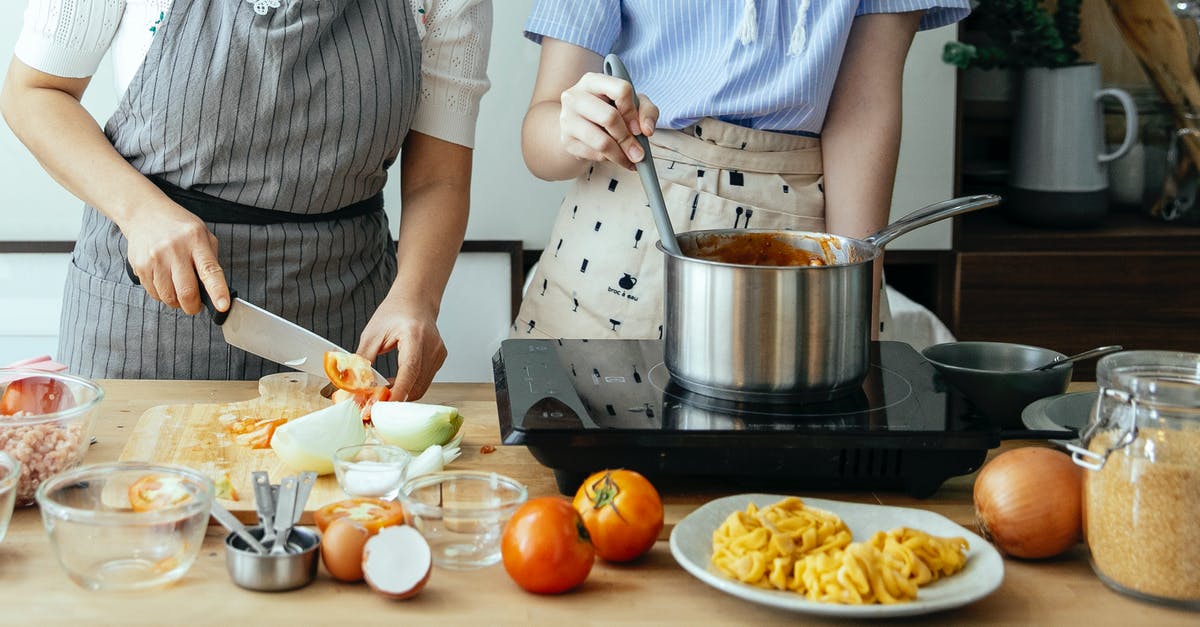 In a tomato sauce recipe, how can I cut the acidity? - Unrecognizable woman cutting fresh tomato on cutting board while standing at table with ingredients and stove near female cook stirring sauce in pan In a tomato sauce recipe, how can I cut the acidity? - Unrecognizable woman cutting fresh tomato on cutting board while standing at table with ingredients and stove near female cook stirring sauce in pan