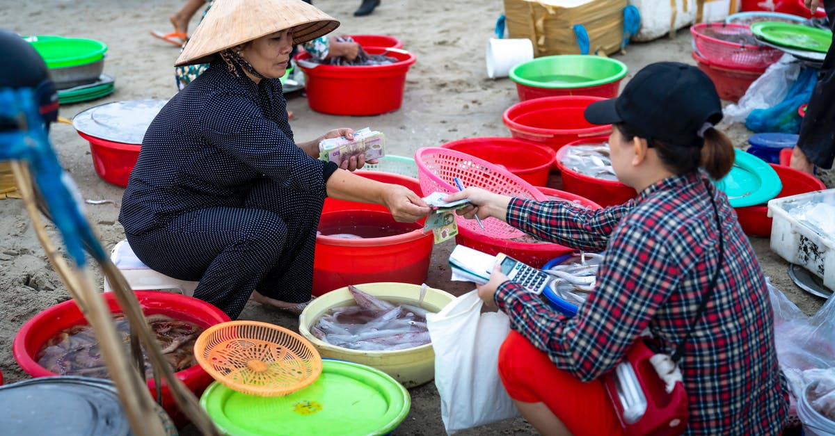 In England, can I buy fresh fish directly off the boat? - Asian female in traditional authentic Vietnamese hat sitting on stool in street fish market and selling seafood to female customer In England, can I buy fresh fish directly off the boat? - Asian female in traditional authentic Vietnamese hat sitting on stool in street fish market and selling seafood to female customer