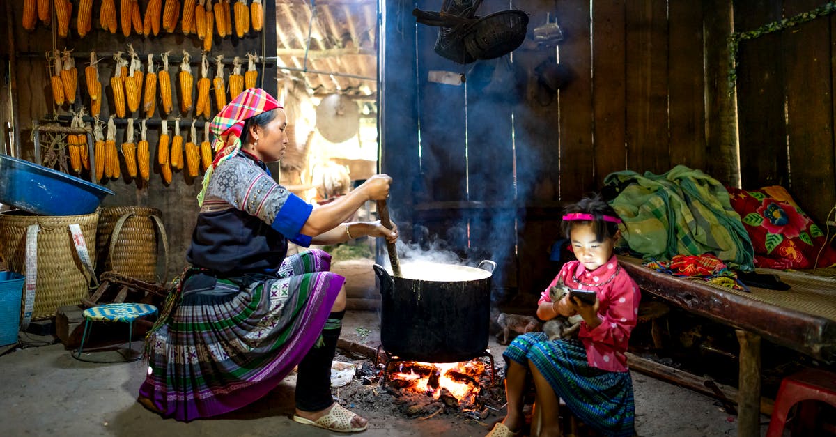 Making Tamales using a corn meal alternative - Side view of Asian woman sitting and preparing food in cauldron against wall with corn while little daughter surfing smartphone in old wooden barn Making Tamales using a corn meal alternative - Side view of Asian woman sitting and preparing food in cauldron against wall with corn while little daughter surfing smartphone in old wooden barn