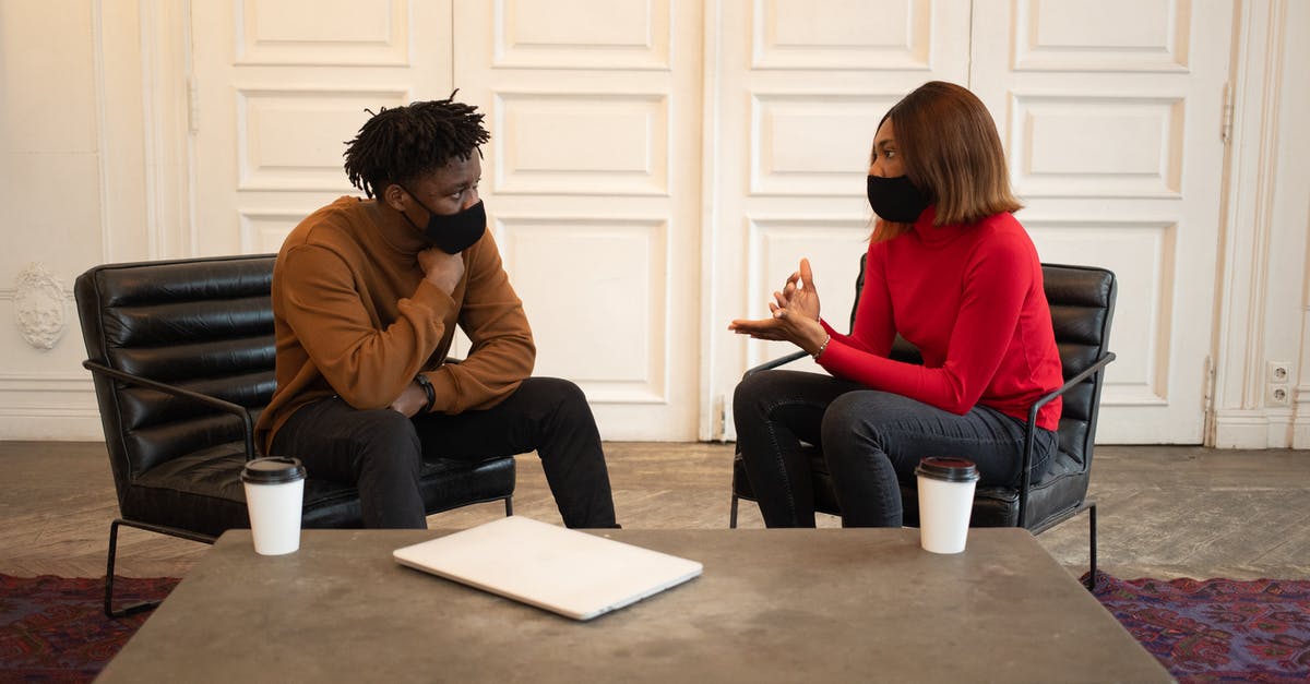 Normal coffee vs decaf - Focused African American colleagues in protective masks looking at each other while having conversation about work at table with laptop and paper cups of takeaway coffee Normal coffee vs decaf - Focused African American colleagues in protective masks looking at each other while having conversation about work at table with laptop and paper cups of takeaway coffee
