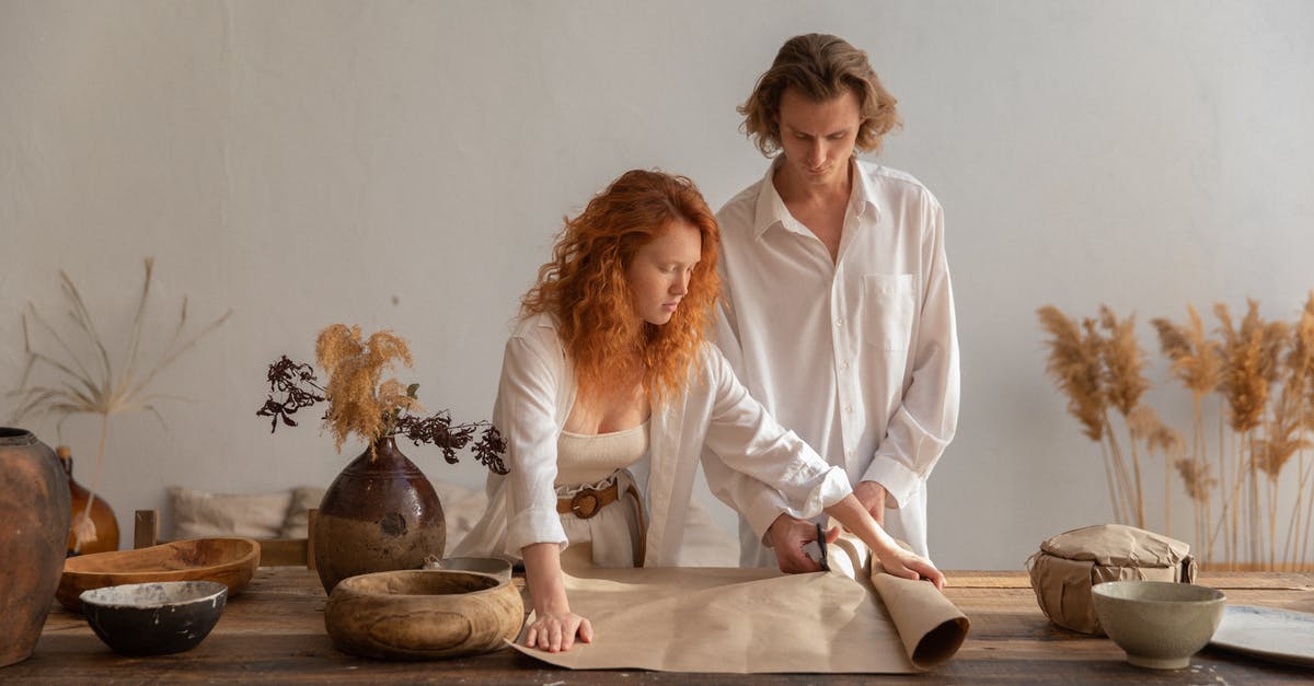On preserving dried ginger - Concentrated man and redhead woman standing near wooden table with craft paper placed near bowl during packaging in cozy workshop On preserving dried ginger - Concentrated man and redhead woman standing near wooden table with craft paper placed near bowl during packaging in cozy workshop