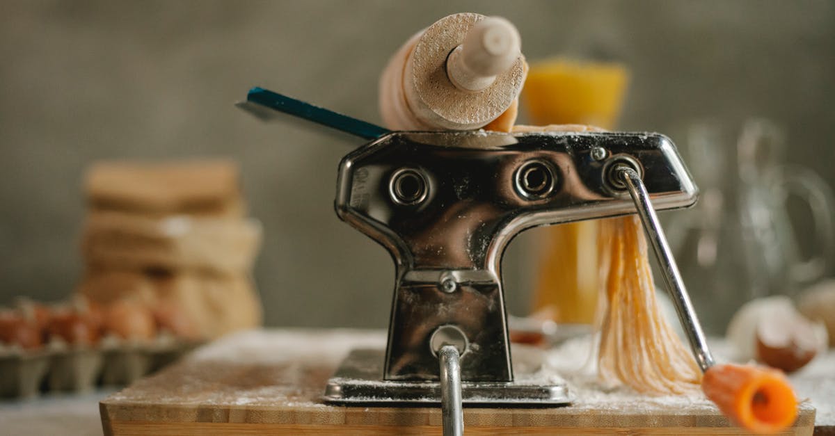 Pasta edges break when being rolled in pasta machine - Stainless modern pasta maker instrument with handle and cut dough placed on table with products on blurred background in kitchen Pasta edges break when being rolled in pasta machine - Stainless modern pasta maker instrument with handle and cut dough placed on table with products on blurred background in kitchen