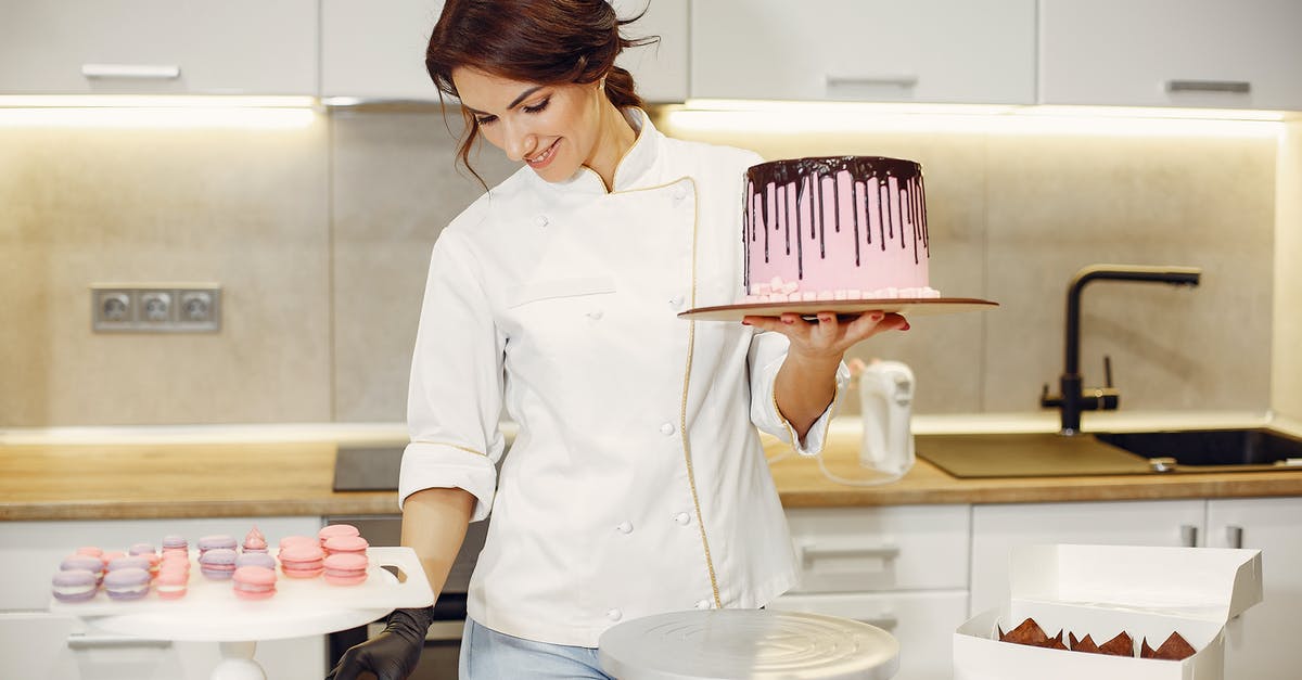 Putting a solid chocolate bar inside cake dough: what's the outcome? - Positive female confectioner in uniform holding pink cake on cardboard with chocolate drips while decorating with colorful marshmallows standing in kitchen Putting a solid chocolate bar inside cake dough: what's the outcome? - Positive female confectioner in uniform holding pink cake on cardboard with chocolate drips while decorating with colorful marshmallows standing in kitchen