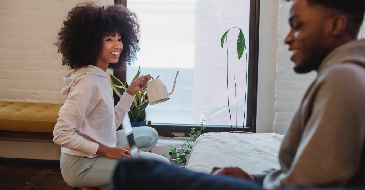 Safe to use pot that has a scratch inside? - Smiling African American woman in casual clothes squatting down and watering plants while spending time with boyfriend browsing laptop at home in daytime Safe to use pot that has a scratch inside? - Smiling African American woman in casual clothes squatting down and watering plants while spending time with boyfriend browsing laptop at home in daytime