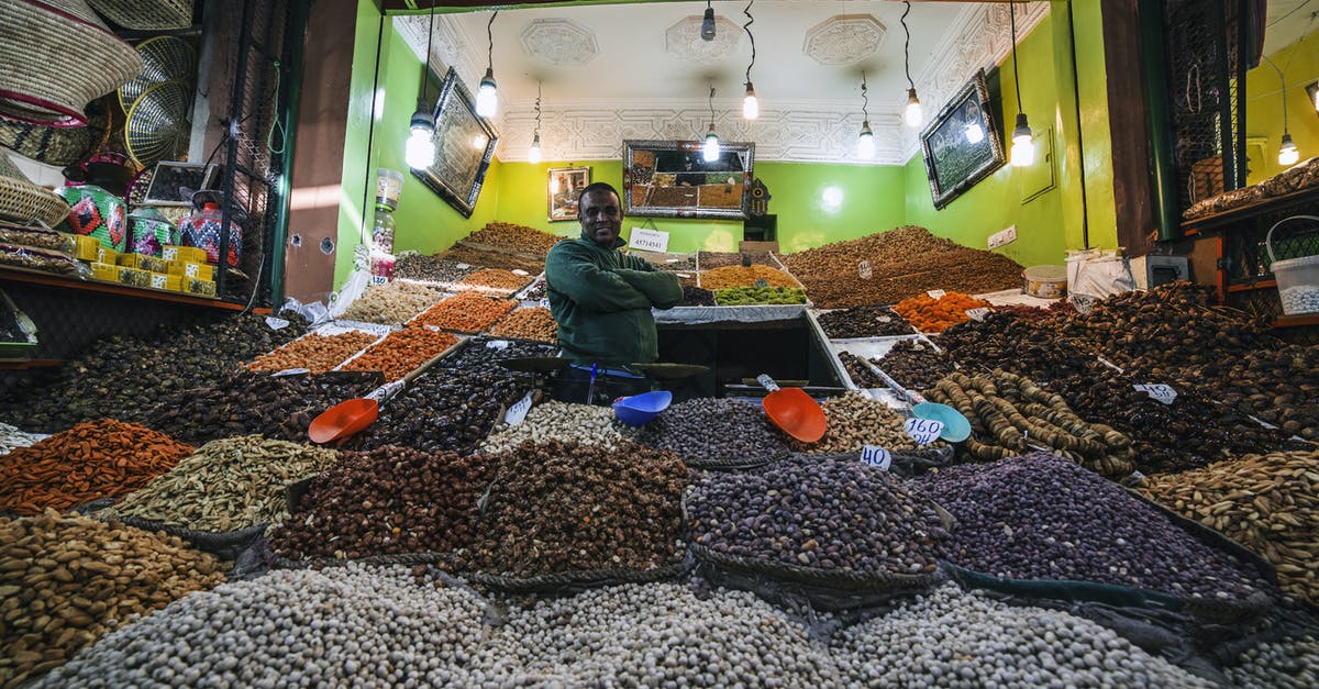 Storing/transporting nut brittle in tins - From below of African American man standing between market stalls with multi colored dried fruits and nuts illuminate with light bulbs Storing/transporting nut brittle in tins - From below of African American man standing between market stalls with multi colored dried fruits and nuts illuminate with light bulbs