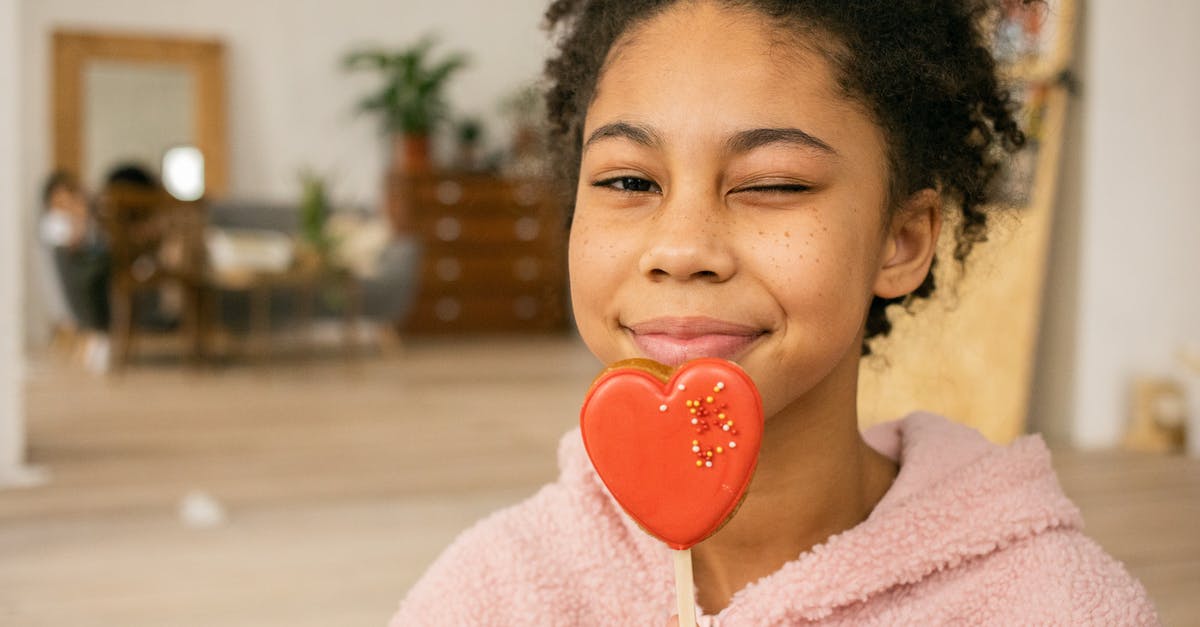 Sugar content of food - Happy African American teenage girl with sweet heart shaped gingerbread cookie on stick winking and looking at camera in light room on blurred background Sugar content of food - Happy African American teenage girl with sweet heart shaped gingerbread cookie on stick winking and looking at camera in light room on blurred background