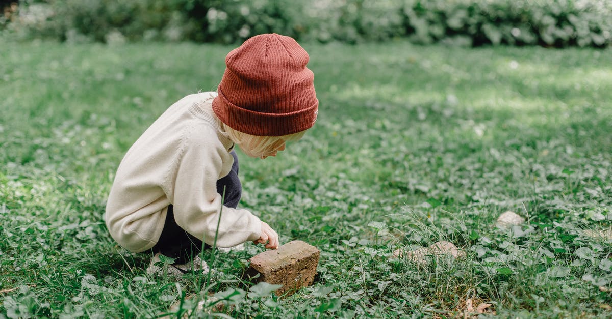 sweet stones of Perosa - Full body side view of playful kid wearing hat squatting on grassy lawn while exploring small stone against blurred background sweet stones of Perosa - Full body side view of playful kid wearing hat squatting on grassy lawn while exploring small stone against blurred background