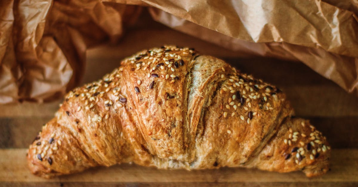 Topping bread with poppy seeds or sesame seeds - Top view of delicious crispy croissant with sesame seeds on wooden table near paper bag Topping bread with poppy seeds or sesame seeds - Top view of delicious crispy croissant with sesame seeds on wooden table near paper bag