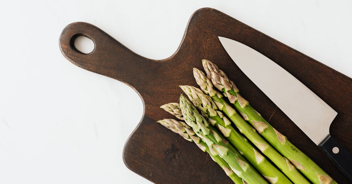 Transfer vegetables objects from cutting board to pan - Top view of bunch fresh green asparagus with knife on dark brown cutting board placed on white surface Transfer vegetables objects from cutting board to pan - Top view of bunch fresh green asparagus with knife on dark brown cutting board placed on white surface