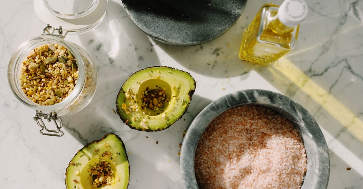 Using avocado oil as a substitute to olive oil - Top view of tasty halved avocado sprinkled with various seeds placed near pink salt with seed mixture in glass pot and other dressing ingredients arranged on white marble table Using avocado oil as a substitute to olive oil - Top view of tasty halved avocado sprinkled with various seeds placed near pink salt with seed mixture in glass pot and other dressing ingredients arranged on white marble table
