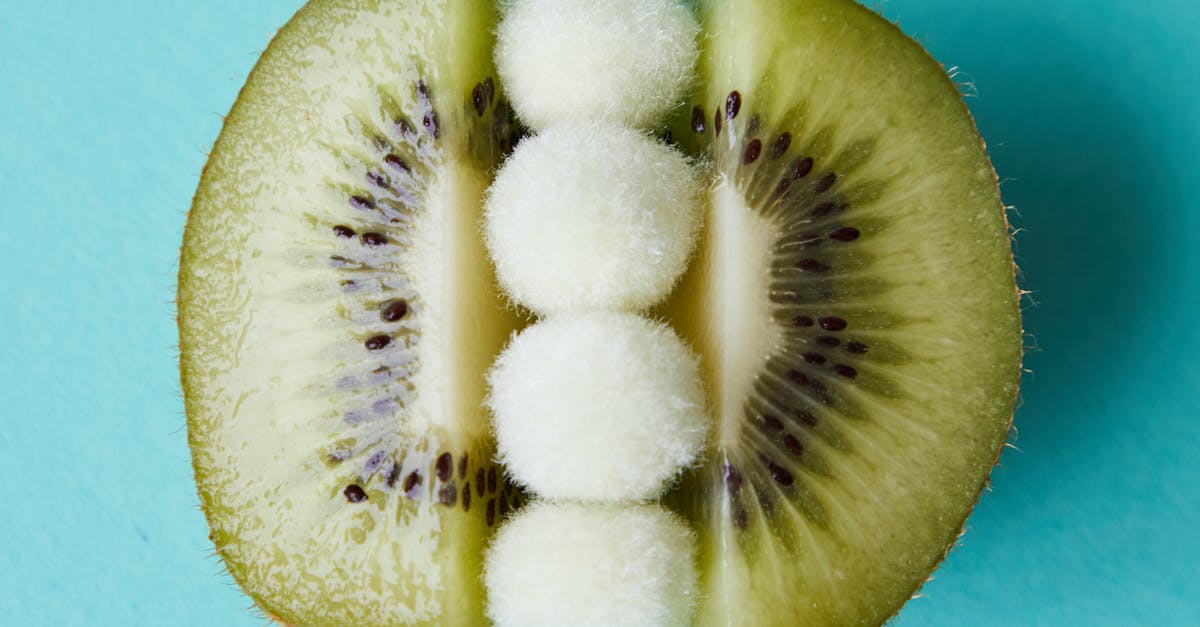 What is this small green fruit called? - Fur pom poms between halves of kiwi on blue surface What is this small green fruit called? - Fur pom poms between halves of kiwi on blue surface