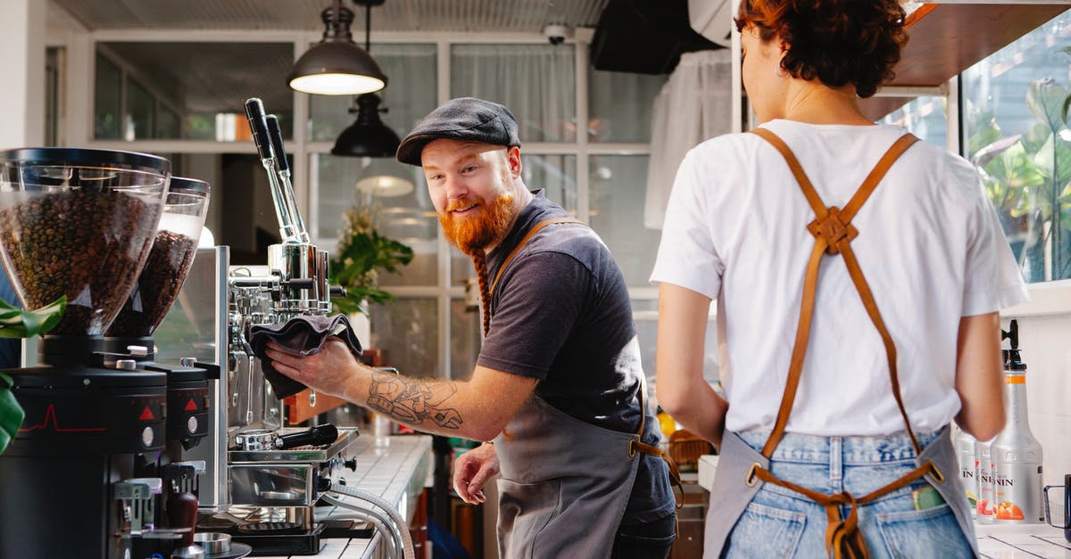 What kind of coffee grinder is best? - Cheerful bearded male cafe employee talking to anonymous female colleague against professional coffee maker and grinders What kind of coffee grinder is best? - Cheerful bearded male cafe employee talking to anonymous female colleague against professional coffee maker and grinders