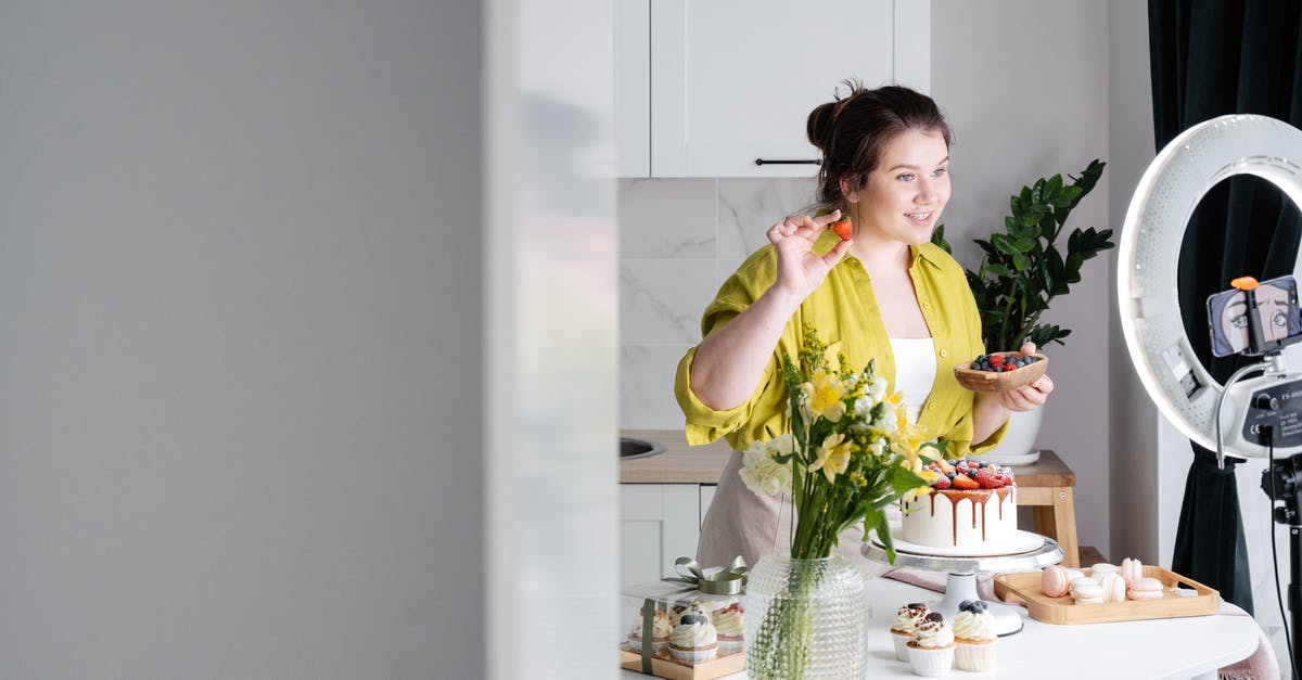 Why is my cupcake brownie like? - Delighted young female influencer in casual clothes smiling and demonstrating fresh berries while decorating appetizing cake during recording vlog on smartphone in kitchen Why is my cupcake brownie like? - Delighted young female influencer in casual clothes smiling and demonstrating fresh berries while decorating appetizing cake during recording vlog on smartphone in kitchen