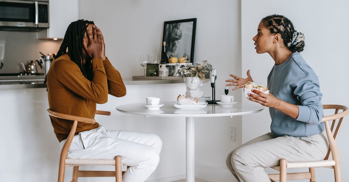 Worried about dangers of rotten food (including smell) - Side view of expressive Hispanic female in casual clothes arguing with African American boyfriend covering face with hands while sitting at table in kitchen at home Worried about dangers of rotten food (including smell) - Side view of expressive Hispanic female in casual clothes arguing with African American boyfriend covering face with hands while sitting at table in kitchen at home